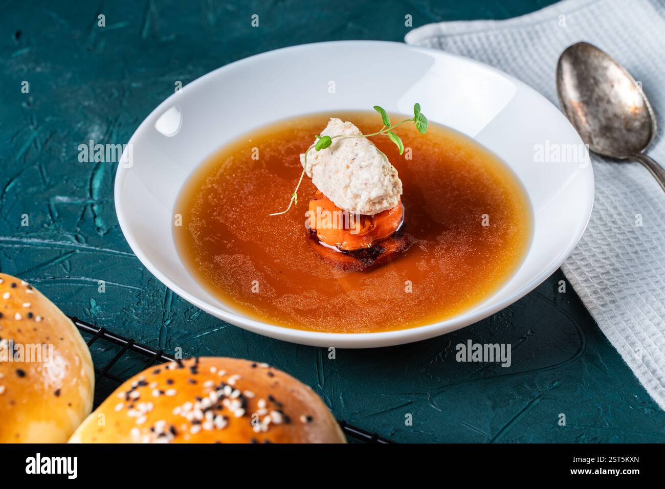 Suppe oder Consomme, dicke Fleischbrühe mit Karotten, Hähnchenknödel serviert auf einem weißen Teller auf dunkelgrünem Hintergrund mit Sesambrötchen, Serviette und Stockfoto