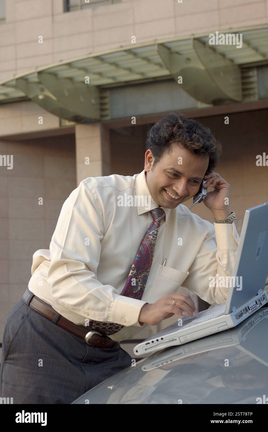 Junger indischer Manager mit Krawatte, lächelnd und telefonierend auf dem Handy und mit Laptop, im Auto vor dem modernen Gebäude Stockfoto