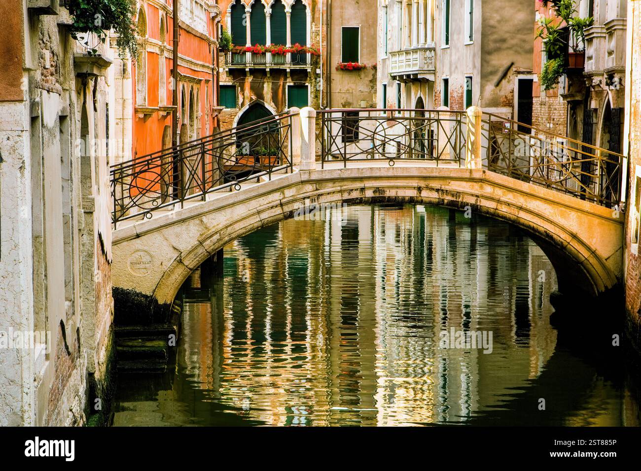 Eine bezaubernde Steinbrücke in Venedig, Italien, führt über einen ruhigen Kanal, flankiert von Gebäuden in warmen Farben mit eleganten Fenstern und Balkonen. Reflexion Stockfoto Eine bezaubernde Steinbrücke in Venedig, Italien, führt über einen ruhigen Kanal, flankiert von Gebäuden in warmen Farben mit eleganten Fenstern und Balkonen. Reflexion Stockfoto