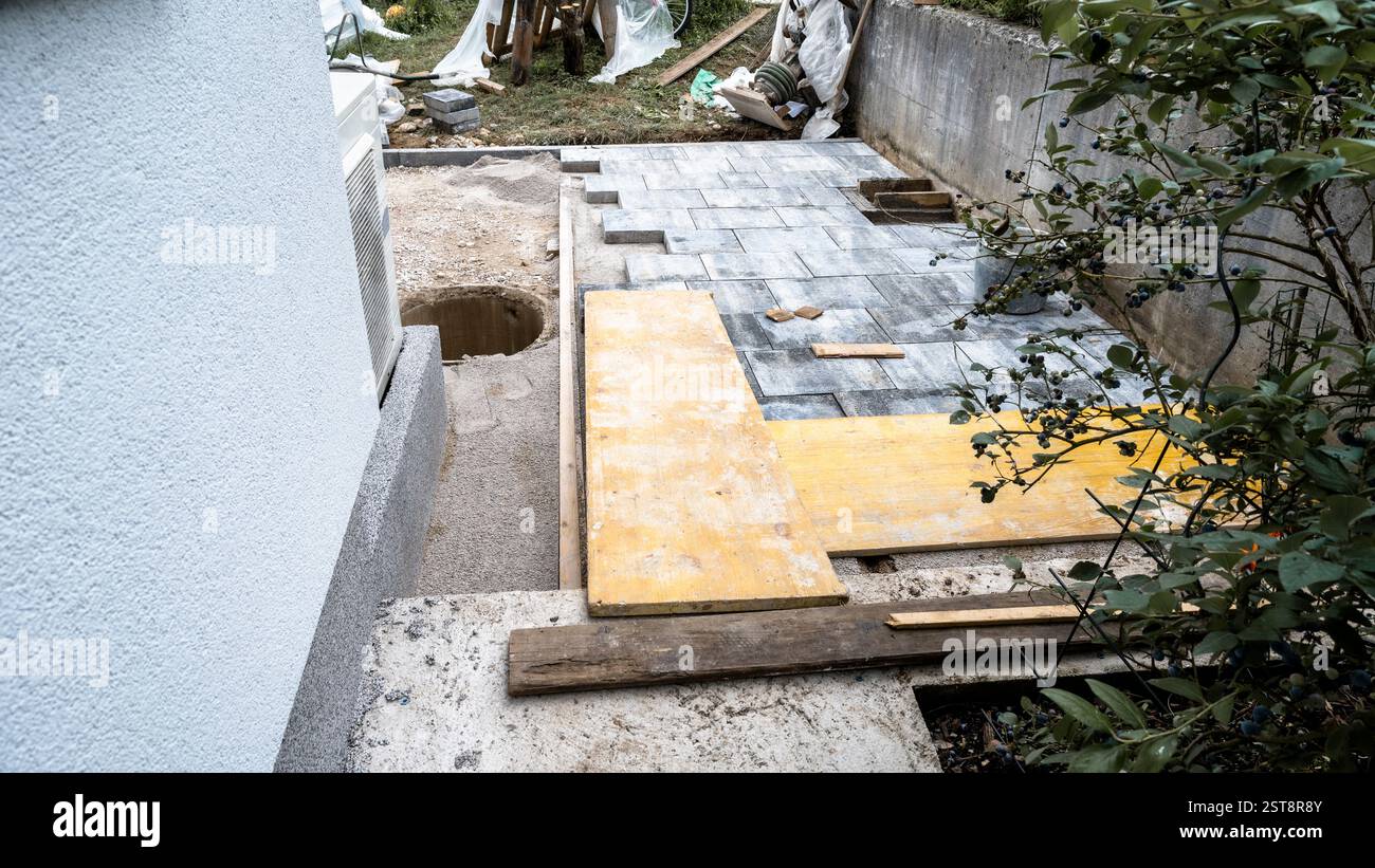 Eine Baustelle mit teilweise fertiggestelltem Steinpflaster, Holzdielen und Werkzeugen, die eine laufende Renovierung des Hinterhofs in der Nähe eines Hauses und eines Blaubeers zeigt Stockfoto