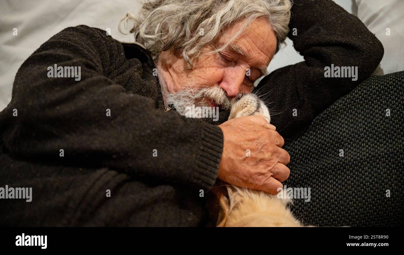 Ein älterer Mann mit lockigen grauen Haaren und Bart umarmt seinen Hund liebevoll und teilt einen zarten Moment der Begleitung und Wärme auf einer gemütlichen Couch. Stockfoto