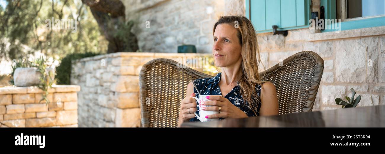 Eine Frau entspannt sich auf einem Korbstuhl, schlürft Kaffee auf einer sonnigen Terrasse mit Steinmauer und Grün und genießt einen ruhigen Moment in einer ruhigen Umgebung im Freien Stockfoto