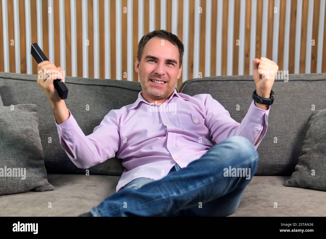 Positiver, fröhlicher Mann, der zu Hause sitzt und Fußballspiele ansieht, entspannen Sie sich drinnen. Der glückliche Kerl auf der Couch schaut mit der Fernbedienung in den Händen fern. Stockfoto