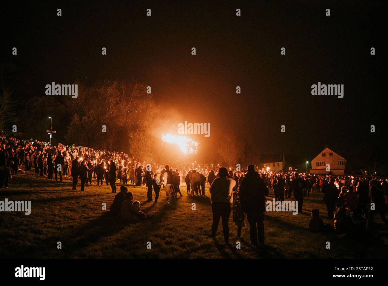 Eine große Menschenmenge versammelt sich auf einem Feld und beobachtet ein Feuer. Die Atmosphäre ist lebhaft und festlich, mit Leuten, die im Gras stehen und sitzen. Stockfoto