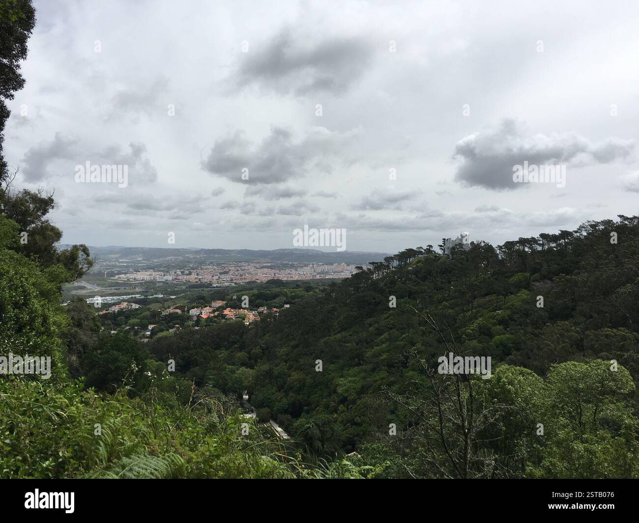Querformat. Üppige grüne Hügel mit dichter Vegetation. Entfernte Stadt mit Gebäuden und bewölktem Himmel. Malerischer Blick auf die Natur und die städtische Entwicklung. Sintra, Po Stockfoto