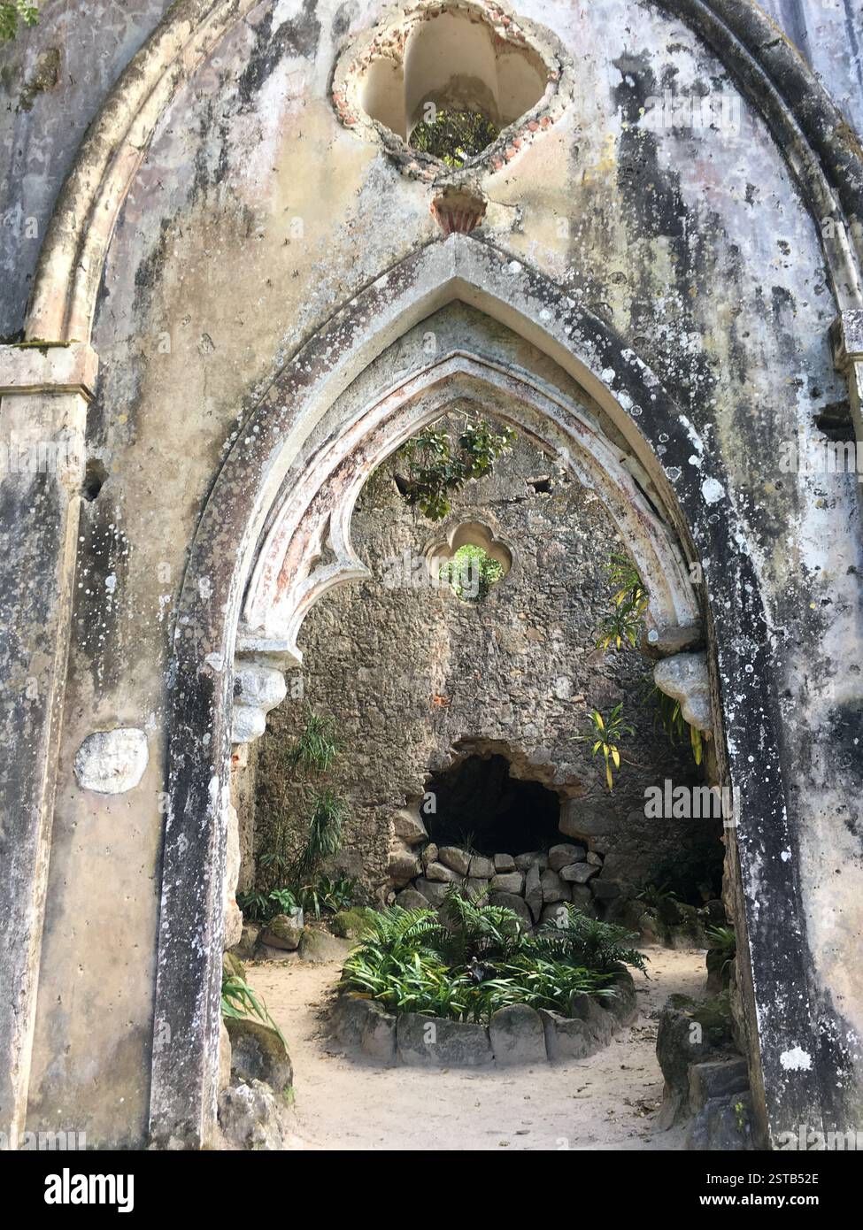 Ruinierter Bogengang in einem üppigen Garten. Moosige Steine und bewachsenes Laub umrahmen den Eingang. Sintra, Portugal. Ein Symbol des Verfalls und der Wiedergeburt, die Natur wiedererlangt Stockfoto