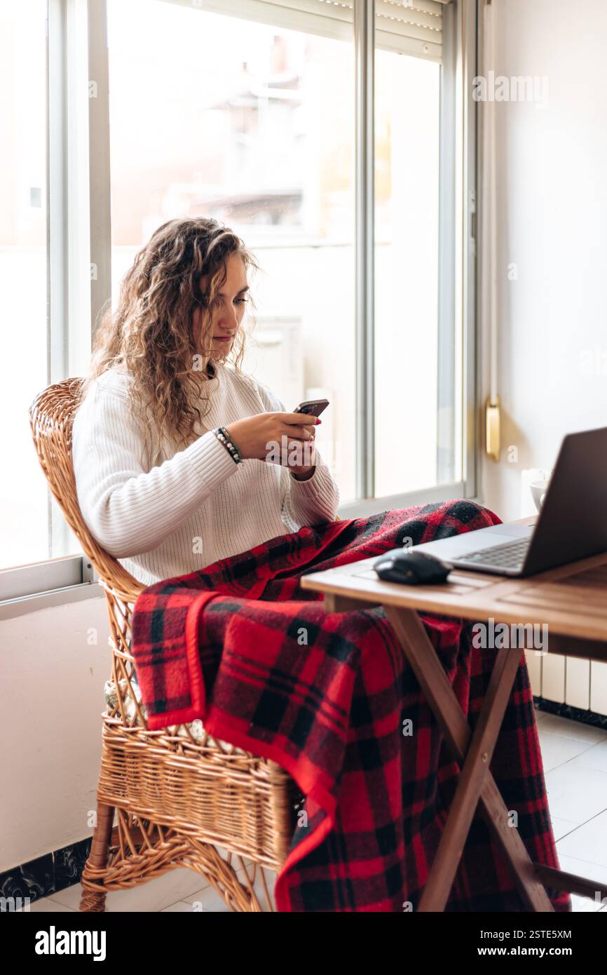 Junge Frau in warmem Pullover, die am Tisch mit Laptop sitzt und das Handy surft, während sie von zu Hause aus arbeitet Stockfoto
