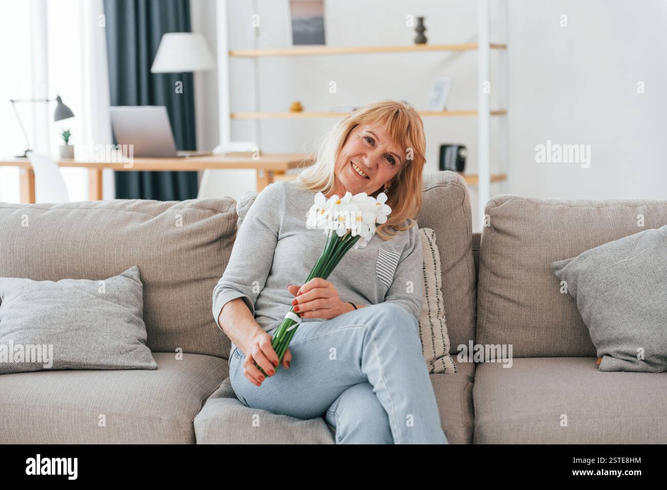 Auf einem Sofa sitzen, Blumen in den Händen. Eine Seniorin mit roten Haaren ist allein zu Hause. Stockfoto
