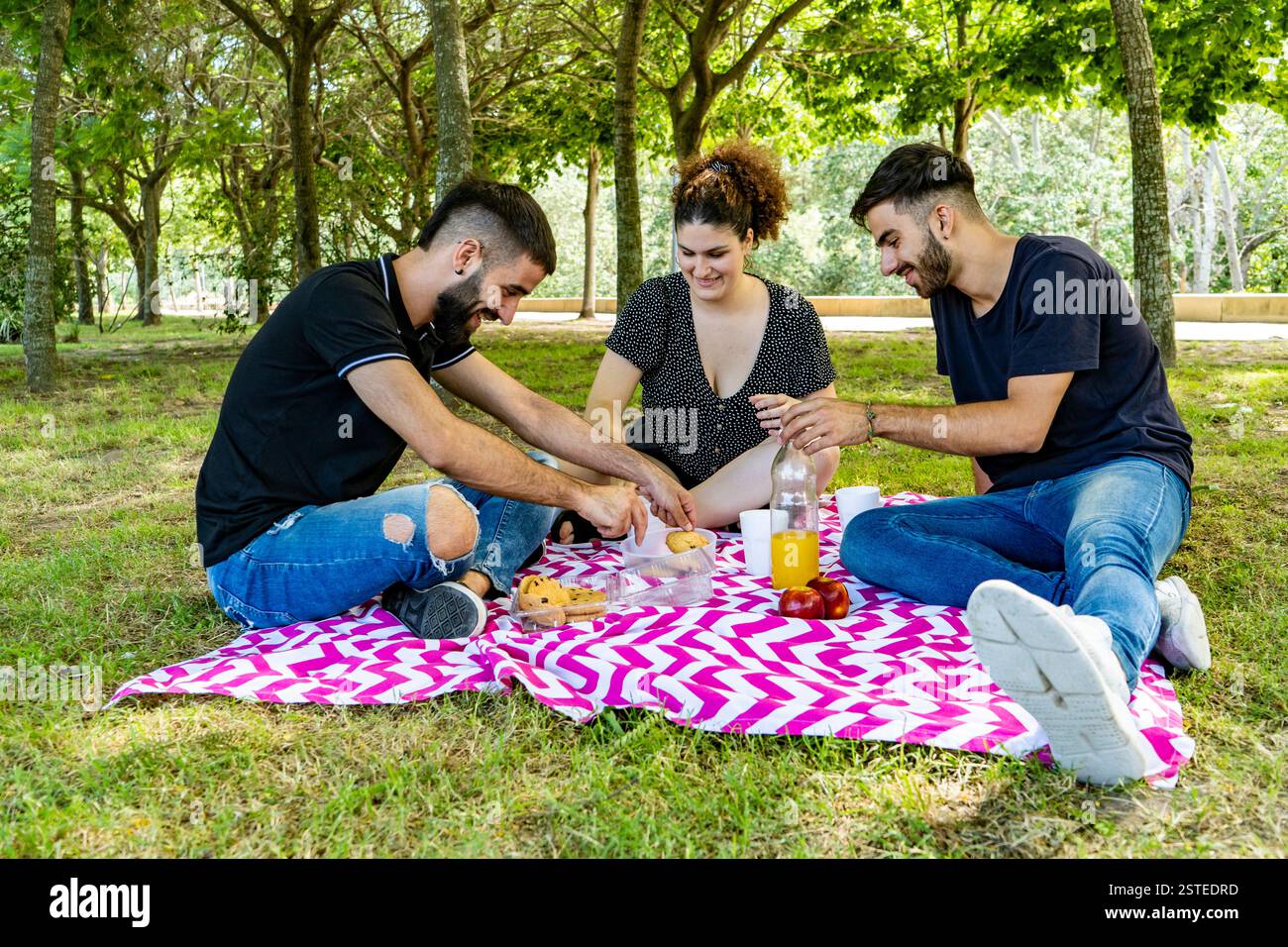 Drei Freunde sitzen auf einer farbenfrohen Picknickdecke in einem üppigen Park, teilen Essen und genießen die Gesellschaft der anderen an einem sonnigen Tag Stockfoto