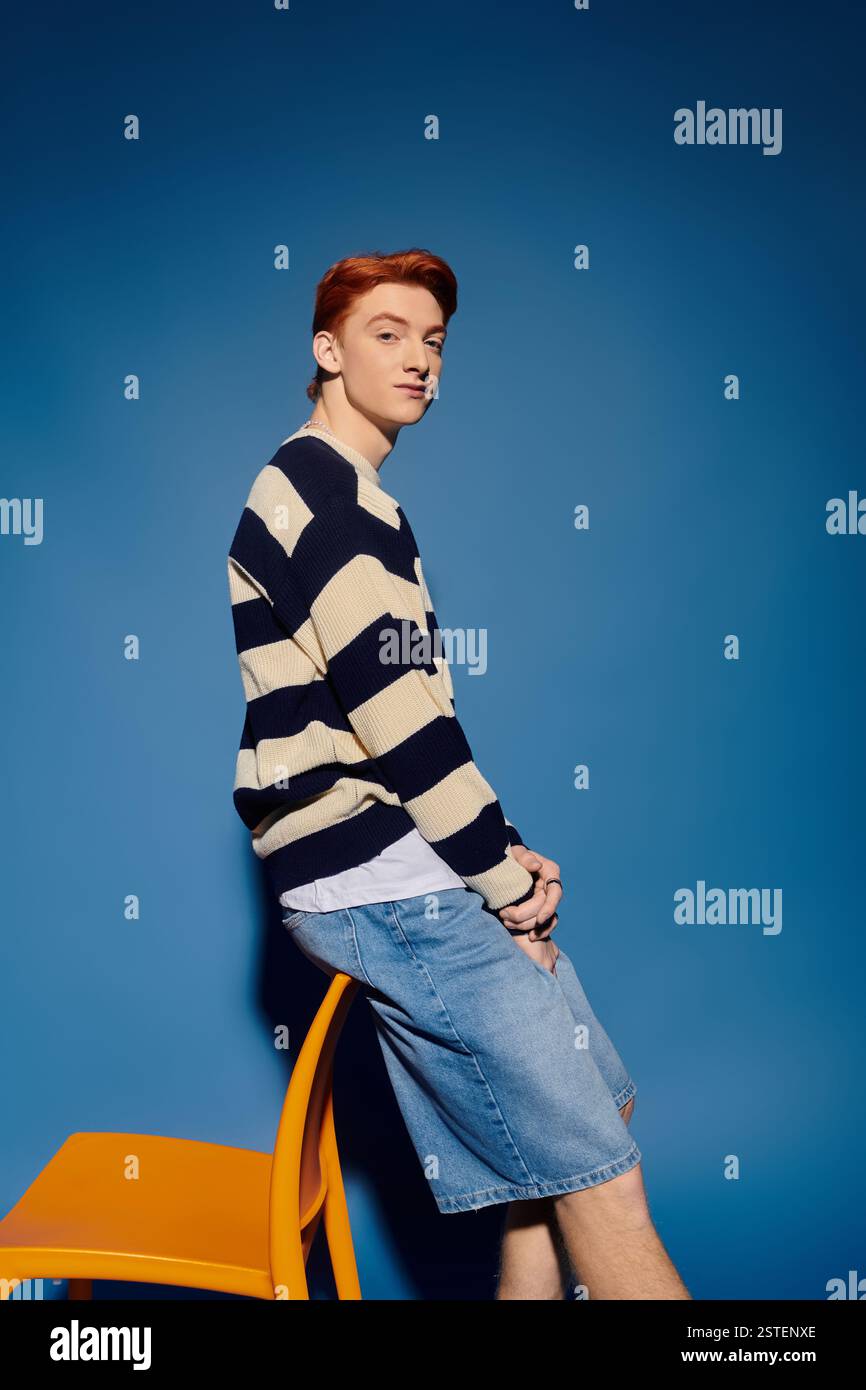Ein junger emotionaler Mann sitzt lässig auf einem Stuhl, verloren in Gedanken, mit einem auffälligen blauen Hintergrund. Stockfoto