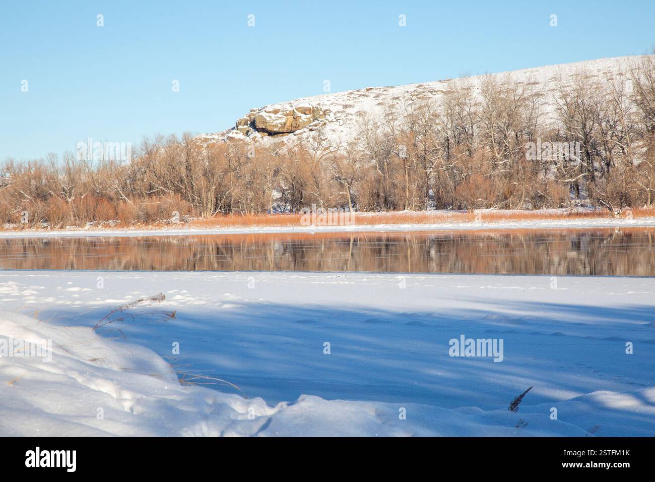 Kühle Winterszene am Missouri River. Felsige Klippen und Pappholzbäume im Hintergrund, Schnee und Eis im Vordergrund. Stockfoto