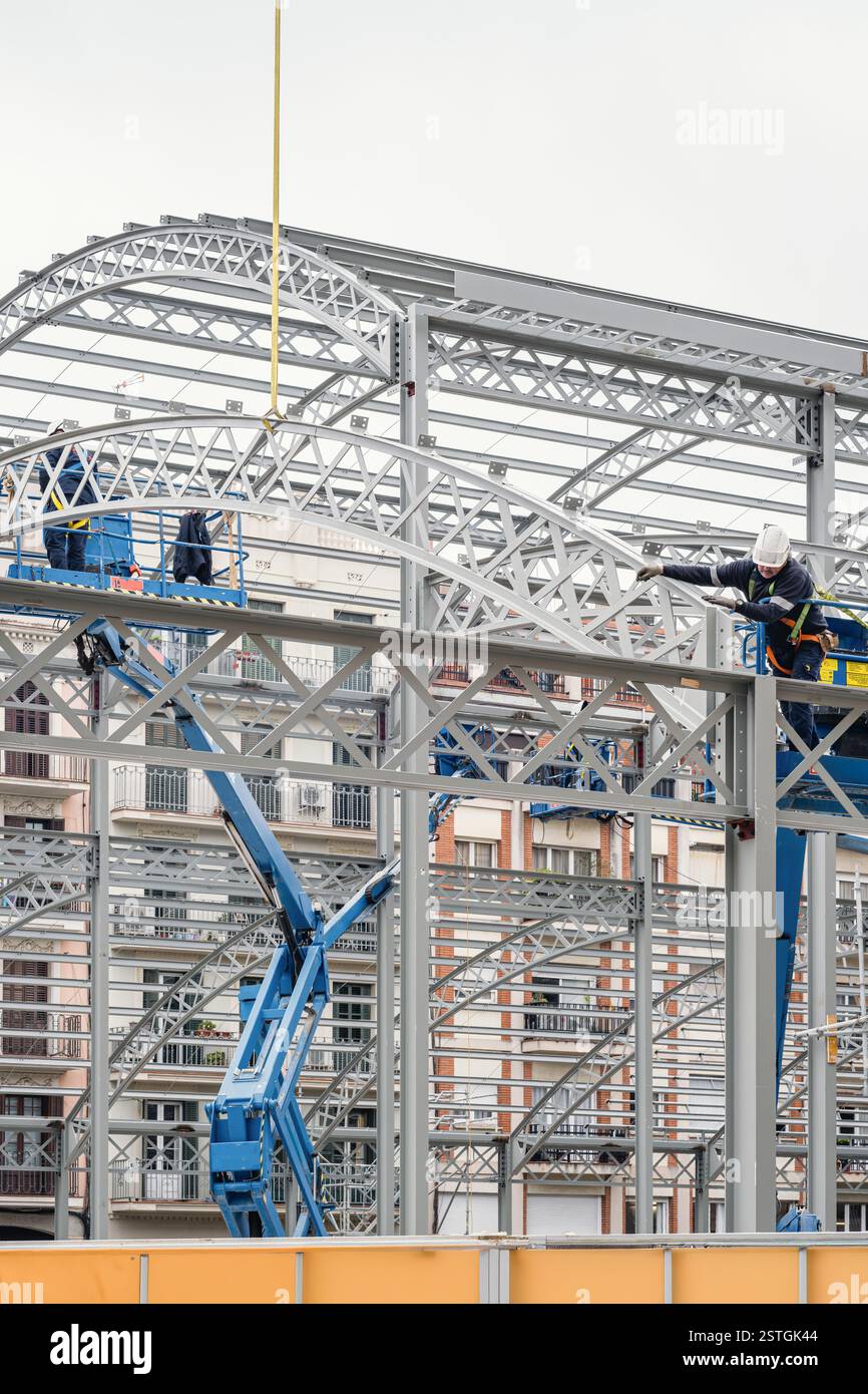 Bauarbeiter, die Hubarbeitsbühnen verwenden, um einen gekrümmten Stahlträger auf einem Dachrahmen zu installieren, die in der Höhe auf einer Baustelle arbeiten. Stockfoto