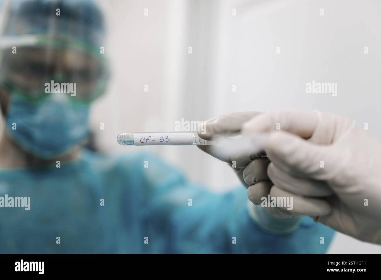 Eine Hand mit Handschuhen, die ein Reagenzglas in einer Laborumgebung hält, mit einer maskierten Person im Blick Stockfoto