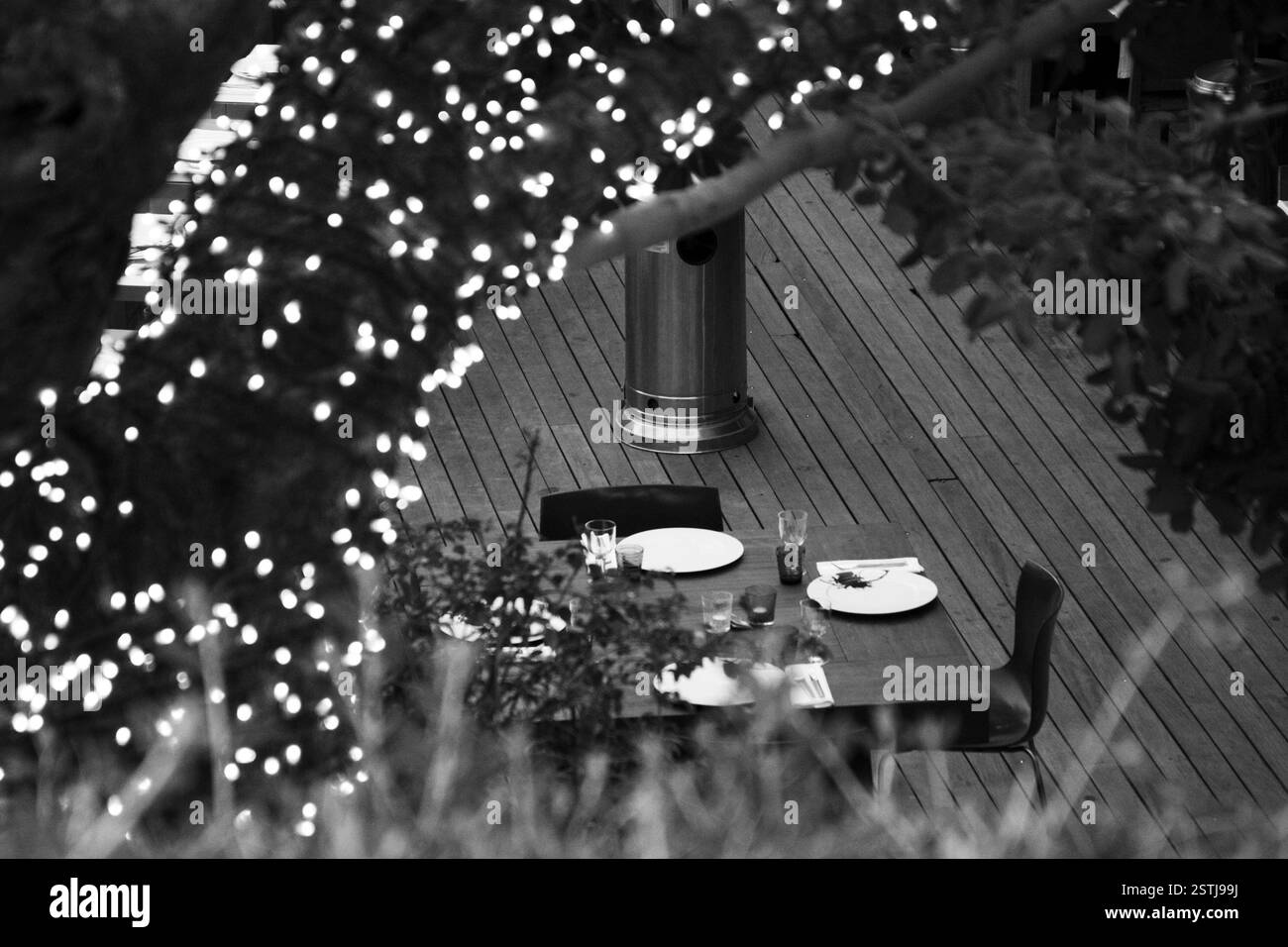 Nahaufnahme eines gedeckten Tisches auf einer Terrasse, mit beleuchteten Zweigen in monochromer Farbe, die ein warmes Ambiente schaffen. Essen im Freien, dekorative Beleuchtung Stockfoto