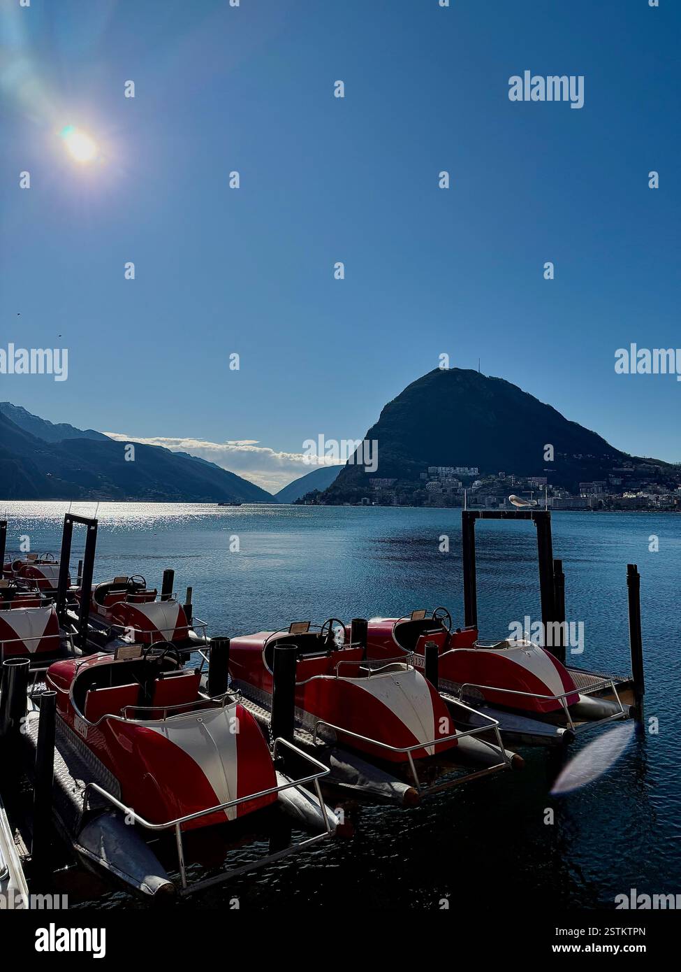 Wunderschöne Landschaft des Sees in Lugano, Schweiz mit kleinen Prahlereien auf einem Dock, blauem Wasser und Bergen im hinteren Teil mit kleinen Gebäuden am Ufer - Smartphone-aufgenommenes Stockfoto