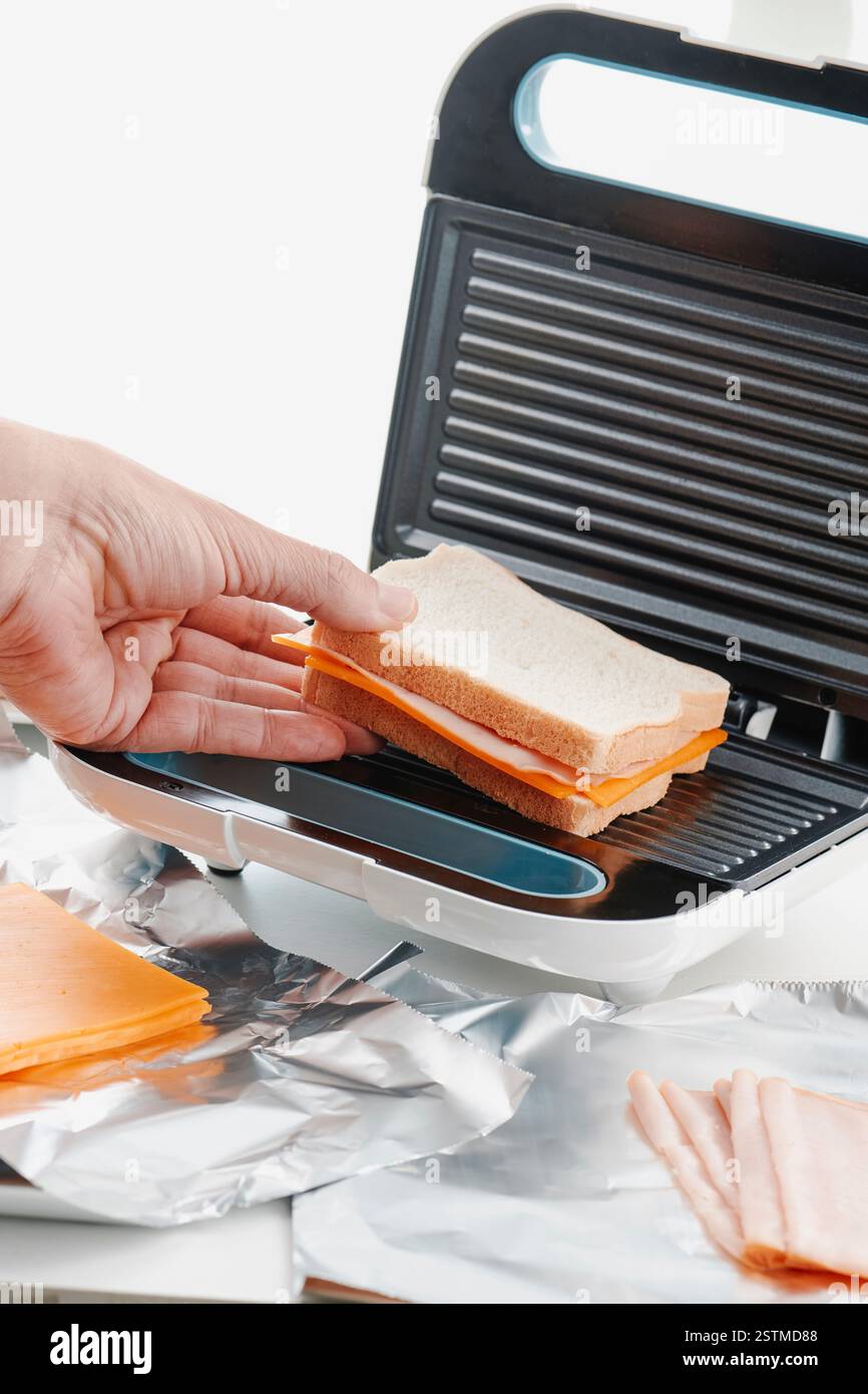 Ein Mann, der ein Schinken-Käse-Sandwich mit einem Sandwich-Toaster zubereitet, auf einer weißen Theke neben einigen Scheiben Cheddar-Käse und Schinken Stockfoto