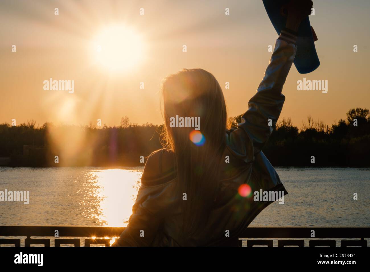 Frau posiert mit den Händen, die das Skateboard in der Höhe des Sonnenuntergangs aufrecht halten, Aufnahme mit Fackeln, niedrigem Kontrast Stockfoto