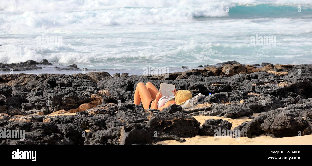 Frau, die Buch liest und sich zwischen vulkanischen Küstenfelsen entspannt, Fuerteventura, Kanarischen Inseln, Spanien, Europa, EU. Aufgenommen Im Dezember 2024 Stockfoto