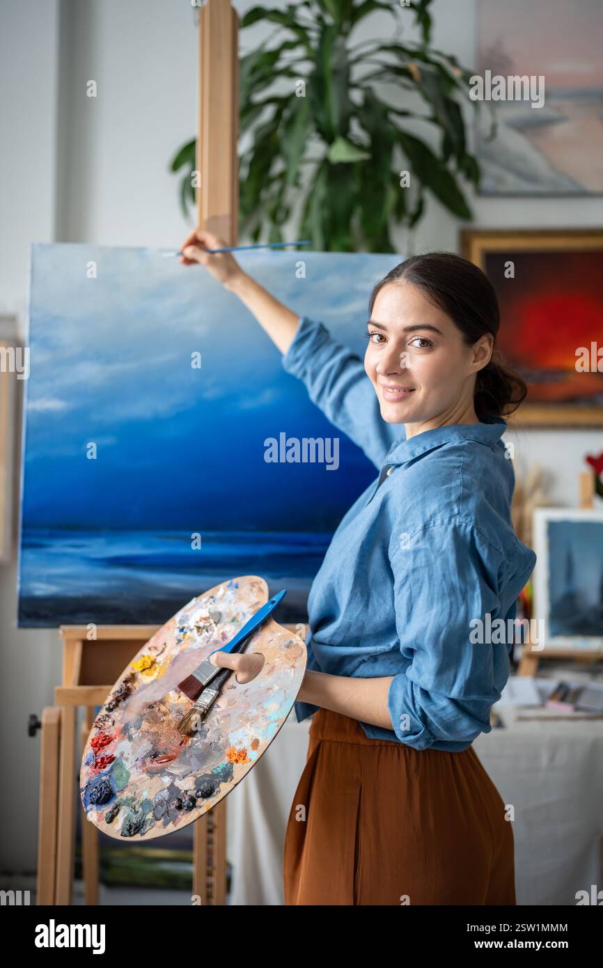 Zufriedene, zufriedene Künstlerin, die an der Malerei arbeitet und Details mit einem kleinen Pinsel zeichnet. Stockfoto