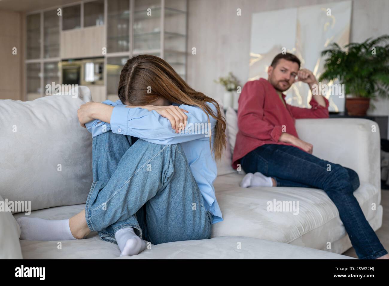 Eine Frau in Tränen, Traurigkeit, versteckt das Gesicht, schrumpft auf dem Sofa, während ihr verzweifelter Ehemann am anderen Ende sitzt Stockfoto Eine Frau in Tränen, Traurigkeit, versteckt das Gesicht, schrumpft auf dem Sofa, während ihr verzweifelter Ehemann am anderen Ende sitzt Stockfoto