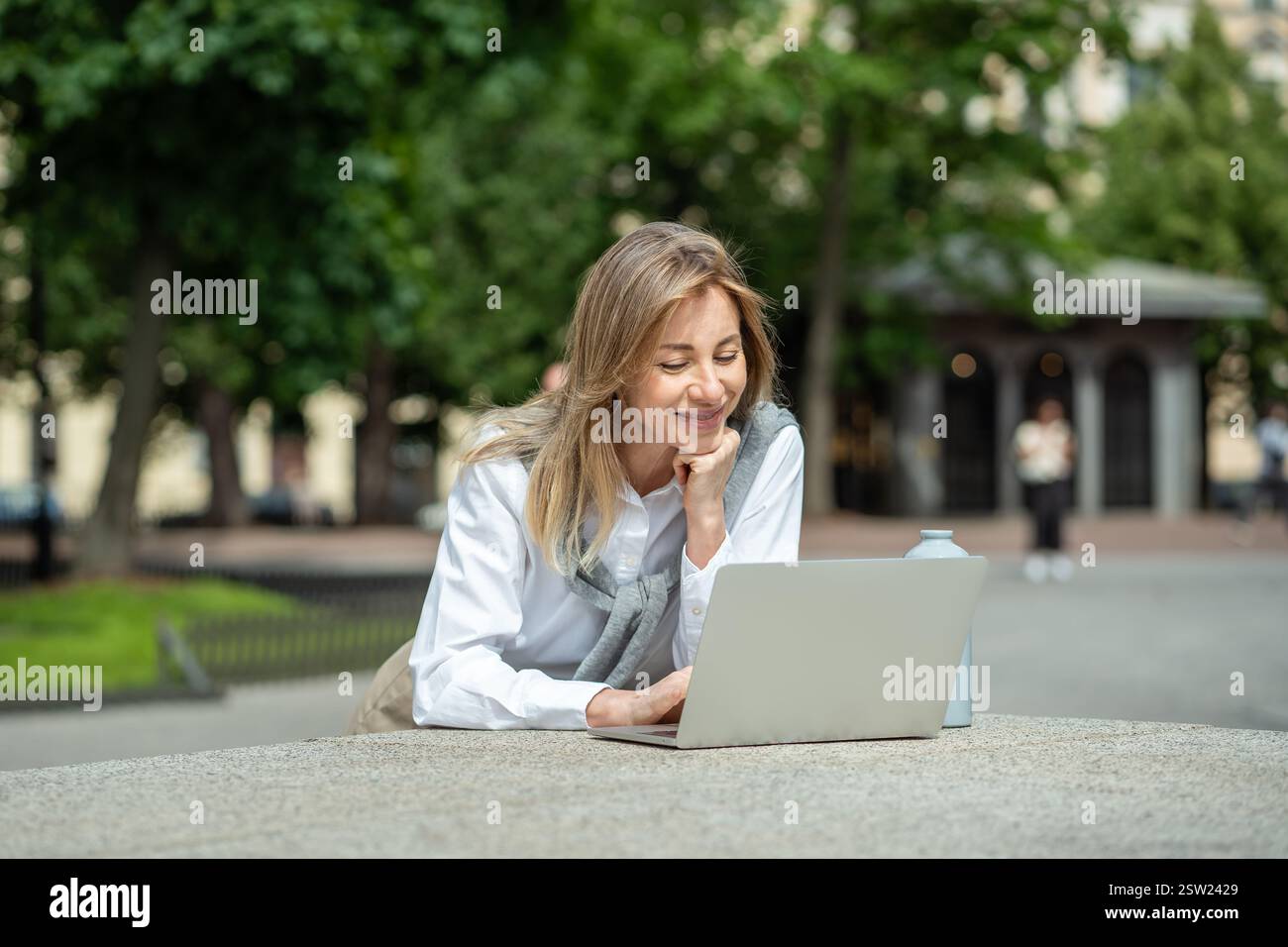 Glückliche Frau, die draußen im Park einen Laptop benutzt und zufrieden lächelt und auf den Bildschirm des Geräts blickt Stockfoto