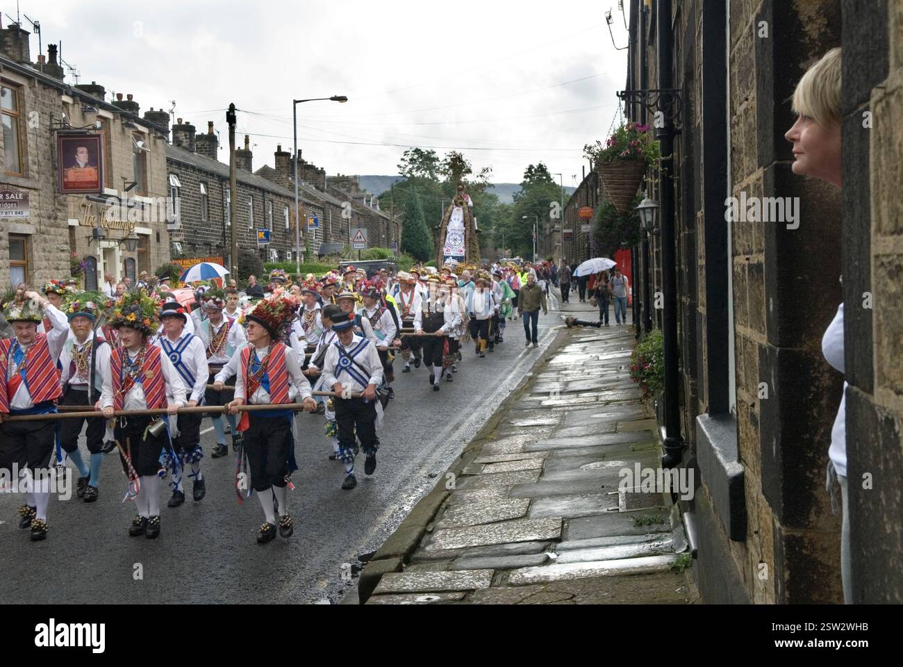Saddleworth Morris Männer und andere Teams von Morris Tänzern ziehen den Rushcart durch die Gemeinde. Eine Frau kommt aus ihrem Terrassenhaus, um die Prozession vorbeiziehen zu sehen. Greenfield, Saddleworth, Lancashire, England 25. August 2012 2010, Großbritannien HOMER SYKES Stockfoto