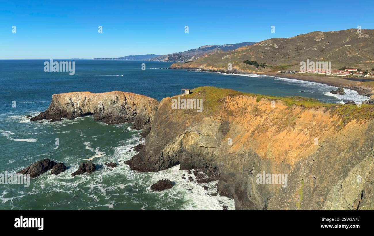 Majestätische Klippen am Point Bonita überragen die stürzenden Meereswellen und erstrecken sich zu einer ruhigen Küste unter einem klaren blauen Himmel. Sanfte Hügel und r Stockfoto