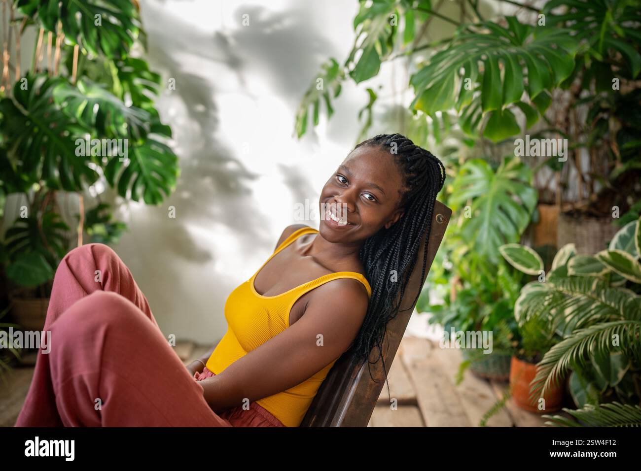 Zufriedene, entspannte schwarze Frau, die die Kamera auf einem gemütlichen Stuhl sieht, umgeben von tropischen Zimmerpflanzen. Stockfoto