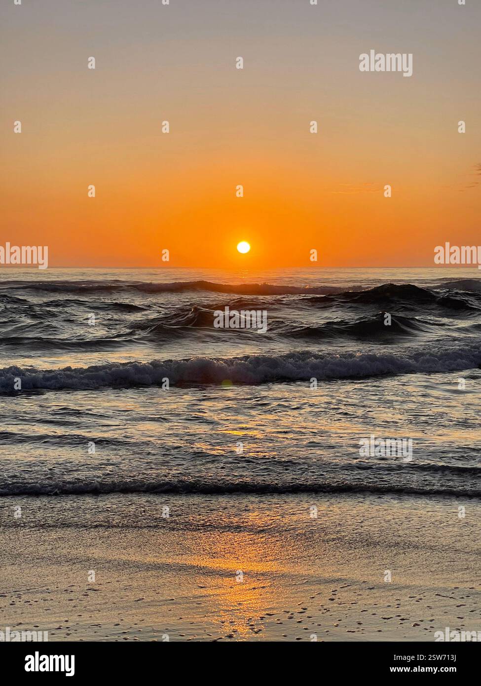 Ein Porträt eines hellen orangen Sonnenaufgangs mit einer Sonne in der Mitte und einer zentralen Reflexion, die über einem Strand mit abgehackten Wellen im Meer aufgeht. Eine Ruhe - Smartphone-aufgenommenes Stockfoto