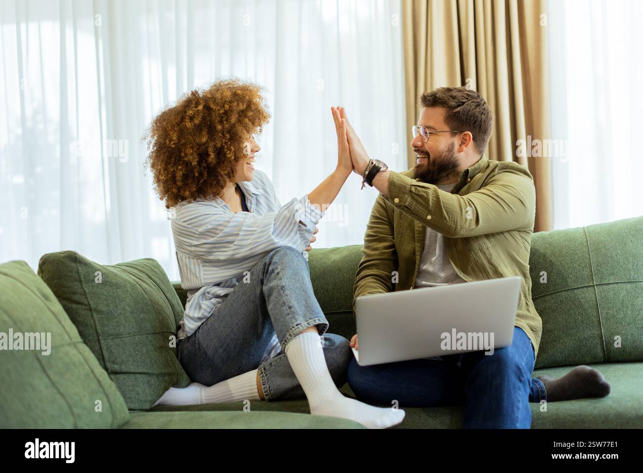 Ein fröhliches Paar sitzt nebeneinander auf einer weichen grünen Couch und führt ein aufregendes Gespräch, während es unter warmem Su auf einem Laptop Ideen durchstöbert Stockfoto