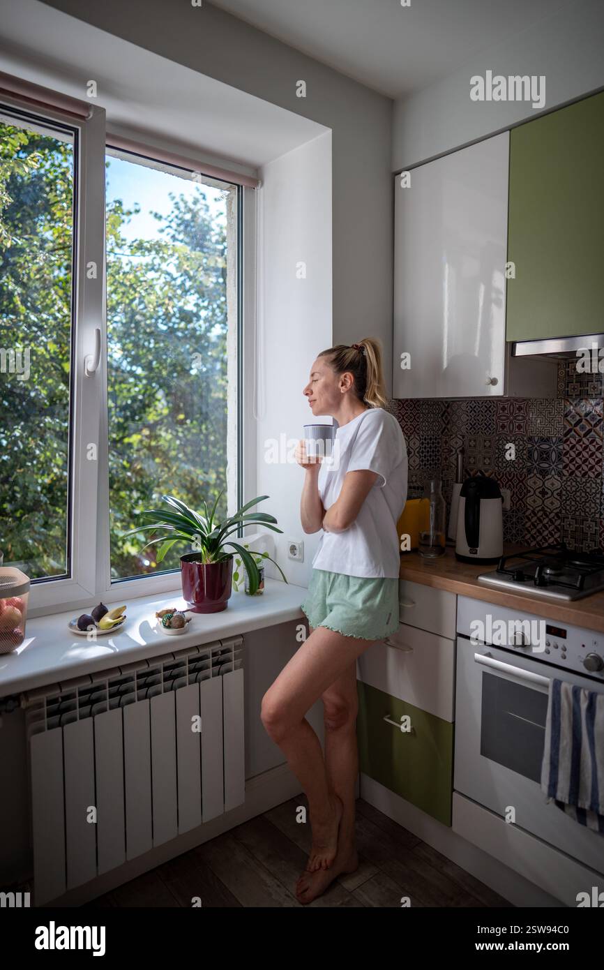 Friedliche Frau schlürft Tee, in Gedanken verloren durch das Küchenfenster. Weibliche reflektierende Momente allein zu Hause Stockfoto