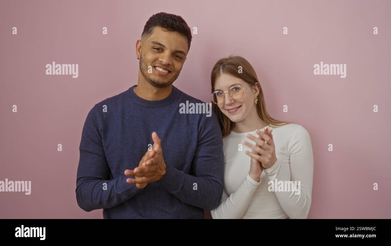 Ein lächelndes Paar, das vor einem rosafarbenen Hintergrund zusammen steht, Mann und Frau glücklich aussehen und mit den Händen klatschen, was Liebe und Verbindung in einem iso-Format repräsentiert Stockfoto