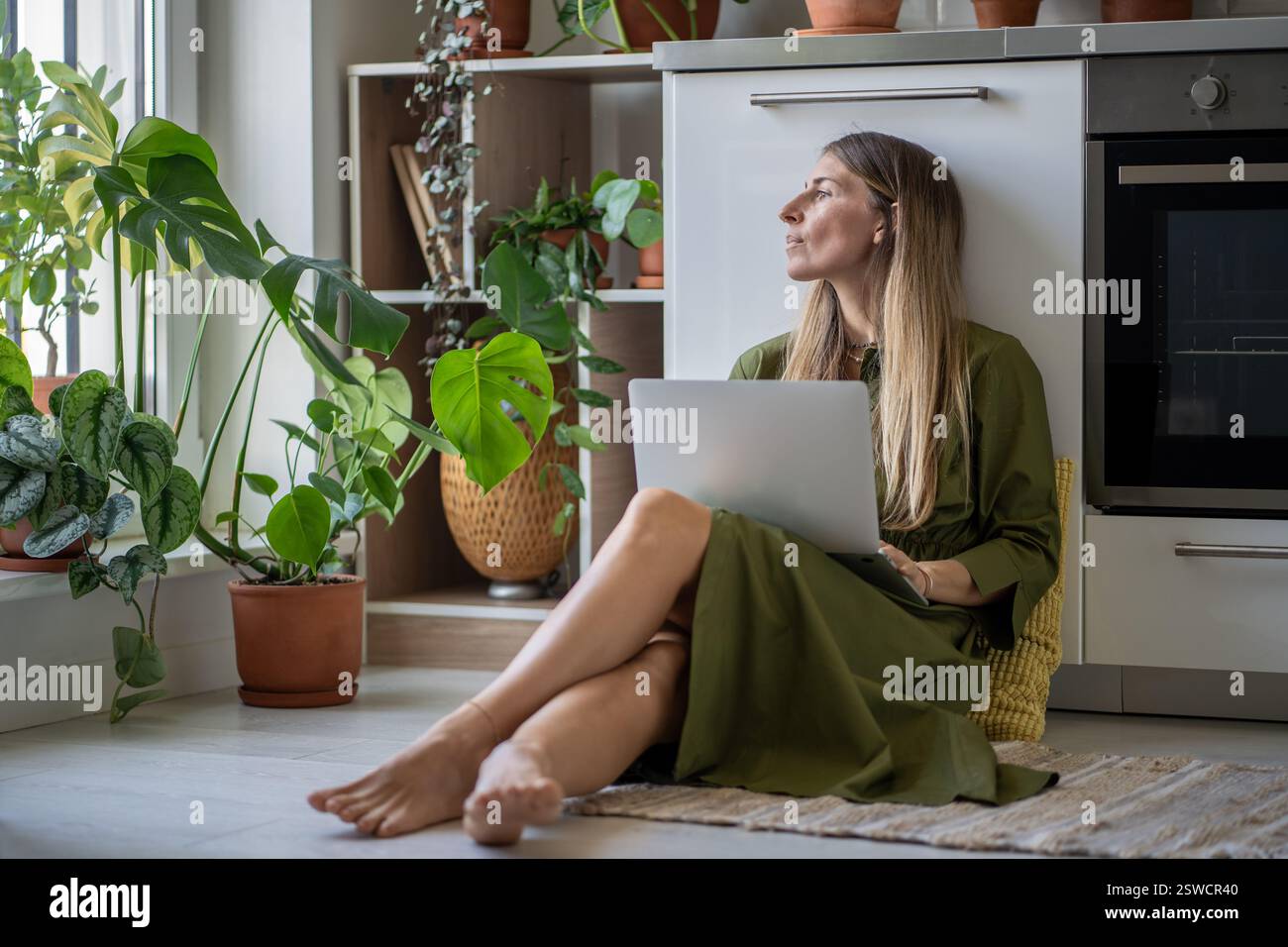 Nachdenkliche Frau mit Laptop, die auf das Fenster blickt, Gefühl von Energiemangel, sitzen zu Hause mit Pflanzen auf dem Boden Stockfoto