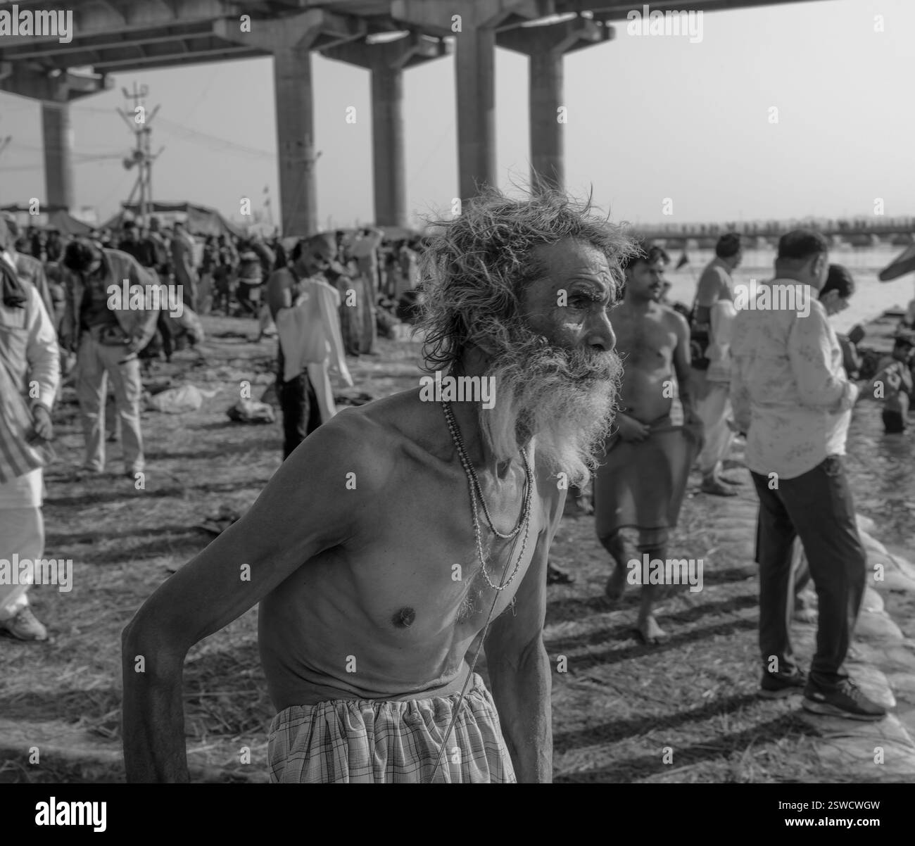 Ein älterer Mann mit markantem Bart betrachtet das geschäftige Ufer während der Maha Kumbh Mela in Prayagraj, Indien, um sich auf ein heiliges Bad vorzubereiten. Stockfoto