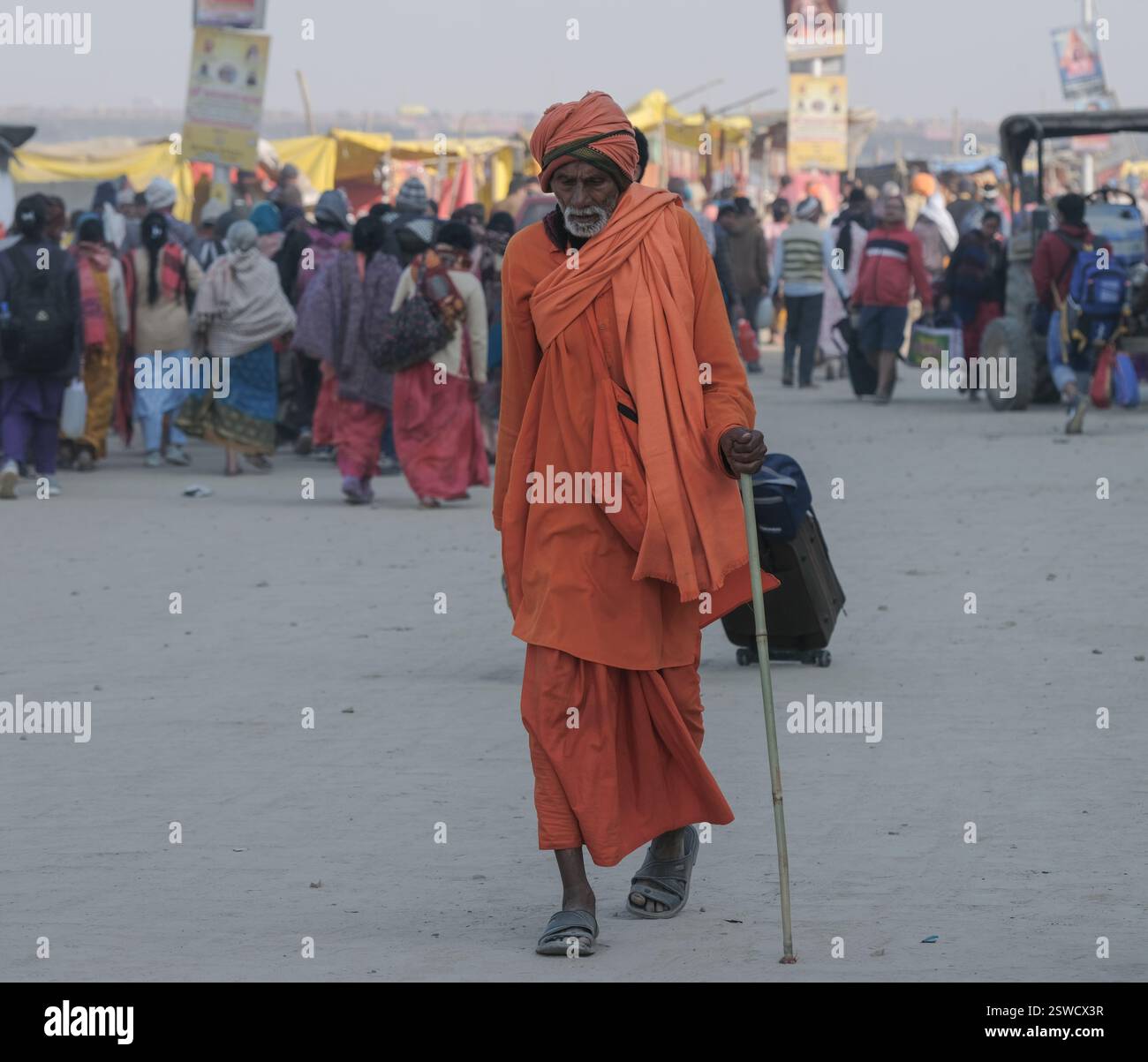 Ein älterer Pilger in orangefarbenen Gewändern spaziert durch die lebhaften Massen von Maha Kumbh Mela in Prayagraj und verkörpert Spiritualität während der Feier. Stockfoto