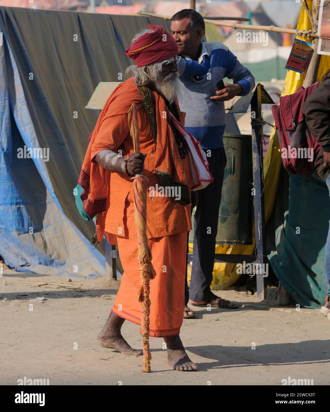Ein älterer Mann in lebendiger traditioneller Kleidung spaziert mit einem Holzpersonal durch die Straße Maha Kumbh Mela in Prayagraj und verkörpert dabei den kulturellen Geist. Stockfoto