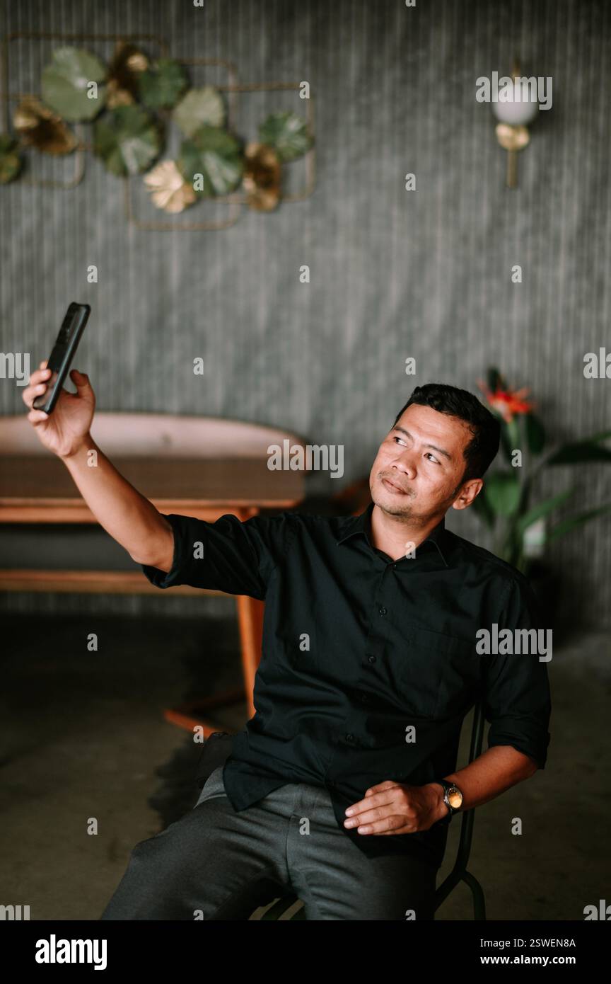 Ein Mann, gekleidet in einem dunklen Hemd und einer grauen Hose, sitzt in einer stilvollen Inneneinrichtung und macht ein Selfie mit seinem Smartphone. Der Hintergrund ist strukturiert mit Stockfoto