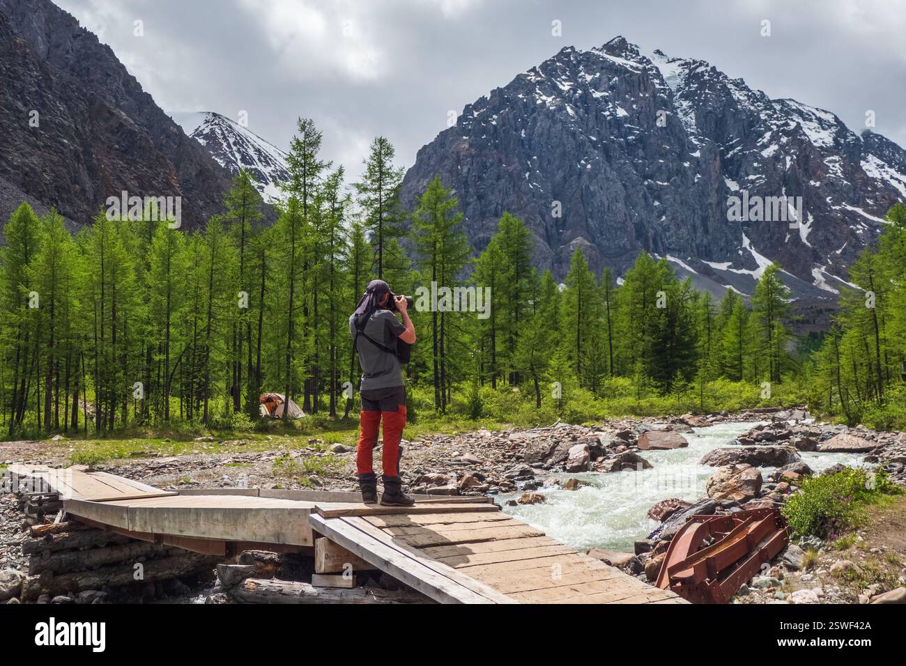 Ein Fotograf auf einer alten Holzbrücke fotografiert ferne Berge. Atemberaubender Blick auf das Bergtal unter dem Blau Stockfoto