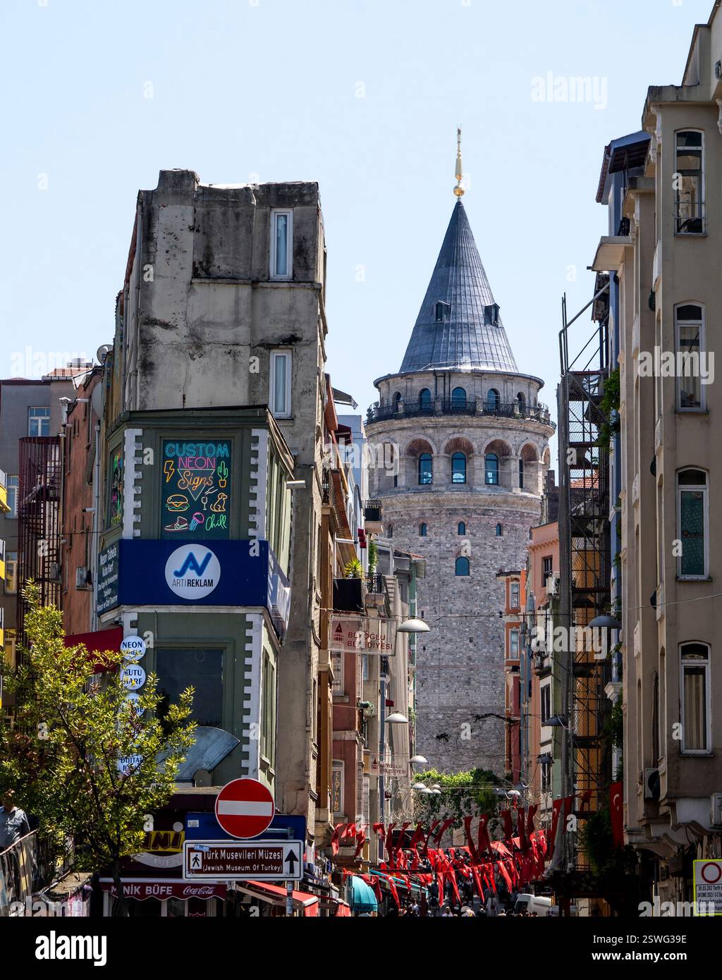 Eine lebendige Straßenszene in Istanbul mit dem berühmten Galatenturm im Hintergrund. Im Vordergrund befinden sich farbenfrohe Gebäude, Straßenschilder und Stockfoto