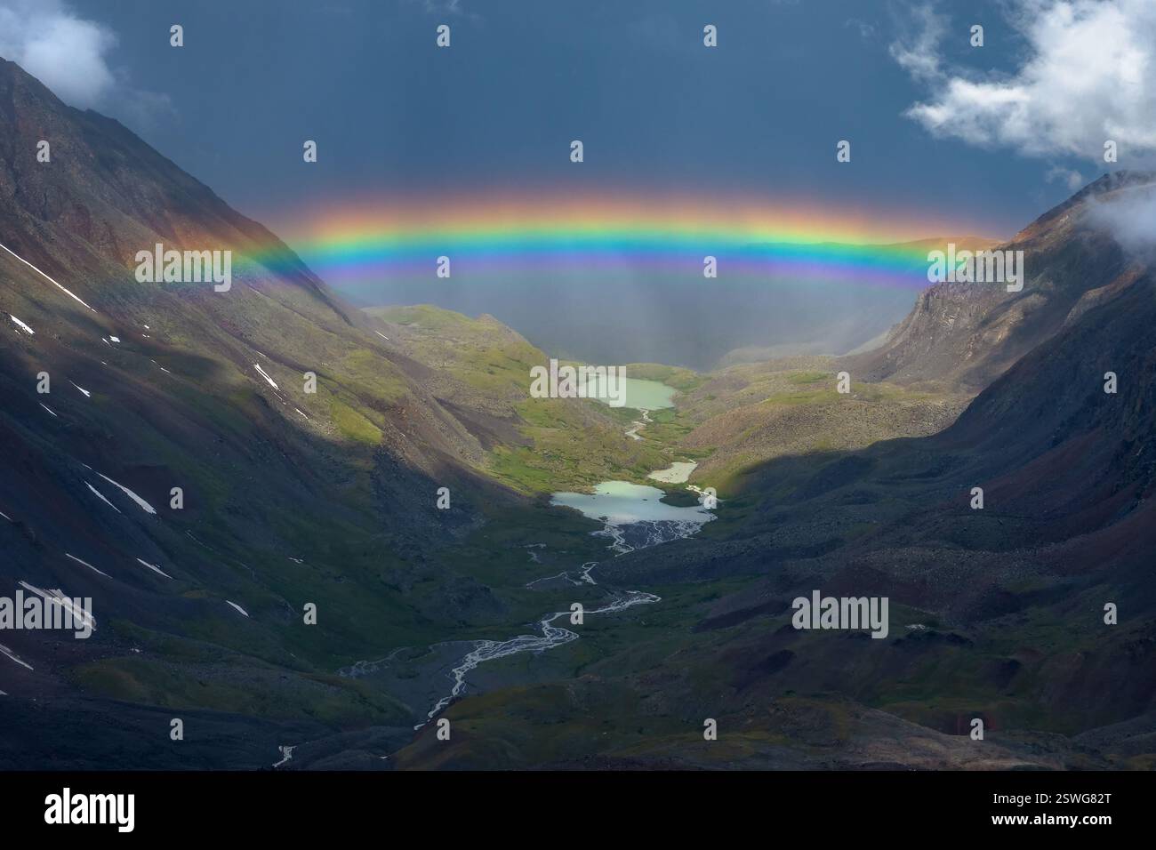 Regenbogen über einem Bergtal. Stimmungsvolle Alpenlandschaft mit schneebedeckten Bergen mit Regenbogen bei regnerischem und sonnigem Wetter. Stockfoto