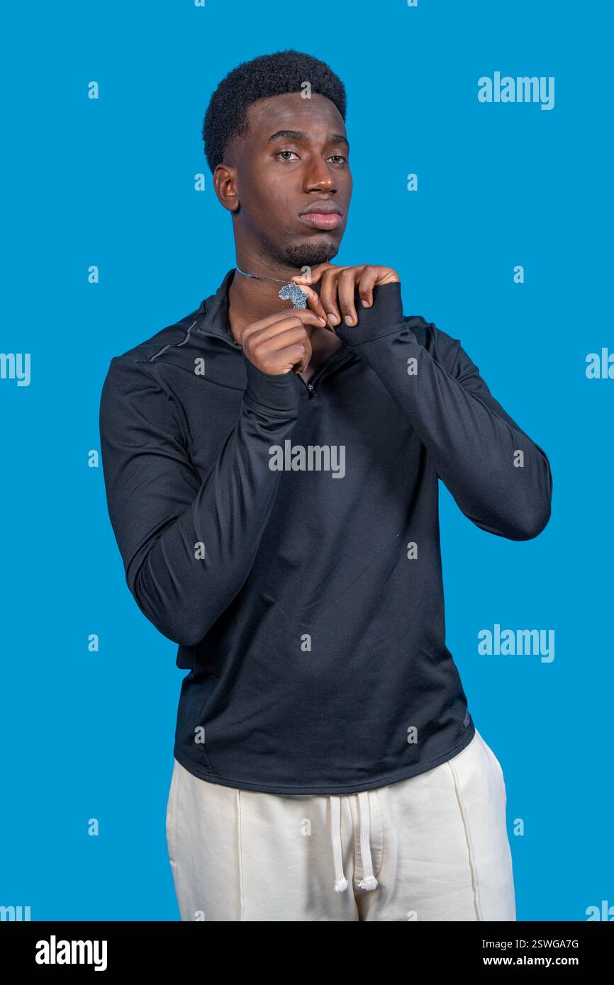 Studio-Porträt eines jungen erwachsenen männlichen Models mit schwarzem Sweatshirt und weißer Hose, der seine Halskette vor einem leuchtend blauen Hintergrund anpasst Stockfoto