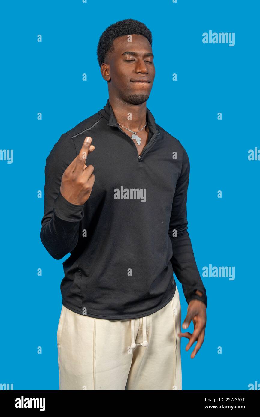 Hoffnungsvoller junger Mann, der die Finger drückt und Glück auf blauem Hintergrund wünscht Stockfoto