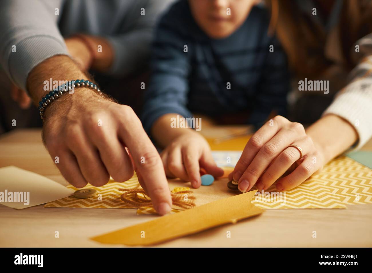 Nahaufnahme von Familienhändlern, die zu Hause an einem DIY-Kunst- und Bastelprojekt zusammenarbeiten, wobei der Schwerpunkt auf der Zusammenstellung verschiedener farbenfroher Materialien liegt. Unscharfer Hintergrund zeigt eine ungezwungene Einstellung an Stockfoto