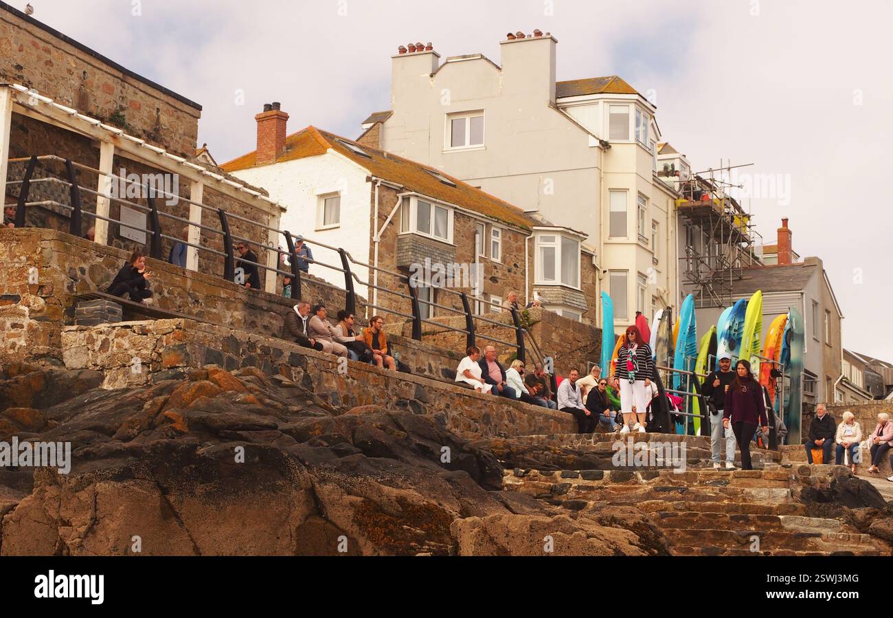 Urlauber sitzen und stehen am New Pier St Ives, Cornwall, England, mit Blick auf das Meer und Sonnenbaden mit Surfbrettern Stockfoto