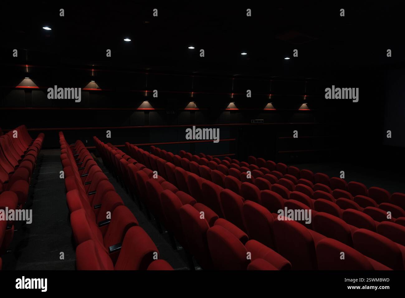 Reihen von leeren roten Sitzen in einem dunklen Theater, beleuchtet von Deckenleuchten, erwarten das Publikum Stockfoto