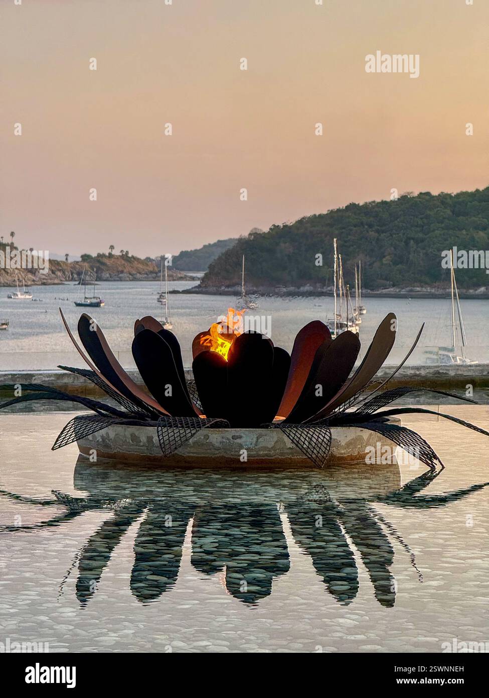 Schwimmende Lotus Fire Pit mit Blick auf eine tropische Bucht Eine Metallfeuerstelle in Form einer Lotusblume schwimmt auf einem ruhigen Wasserbecken, mit Flammen, die darauf leuchten Stockfoto