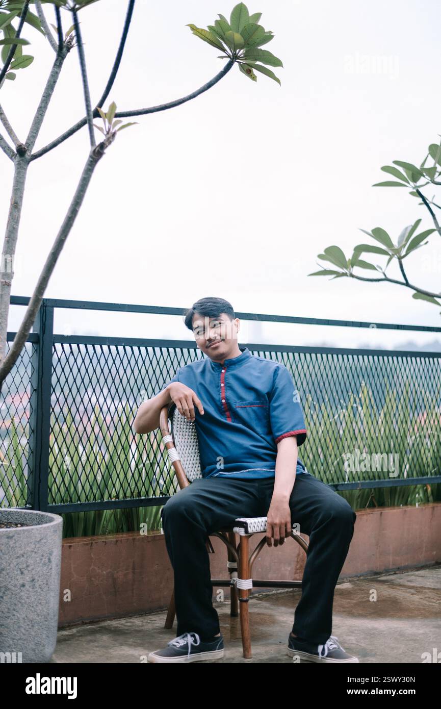Ein junger Mann in lässiger Kleidung sitzt bequem auf einem Stuhl auf einer Dachterrasse. Er lächelt sanft und scheint entspannt zu sein und genießt einen ruhigen Moment Stockfoto