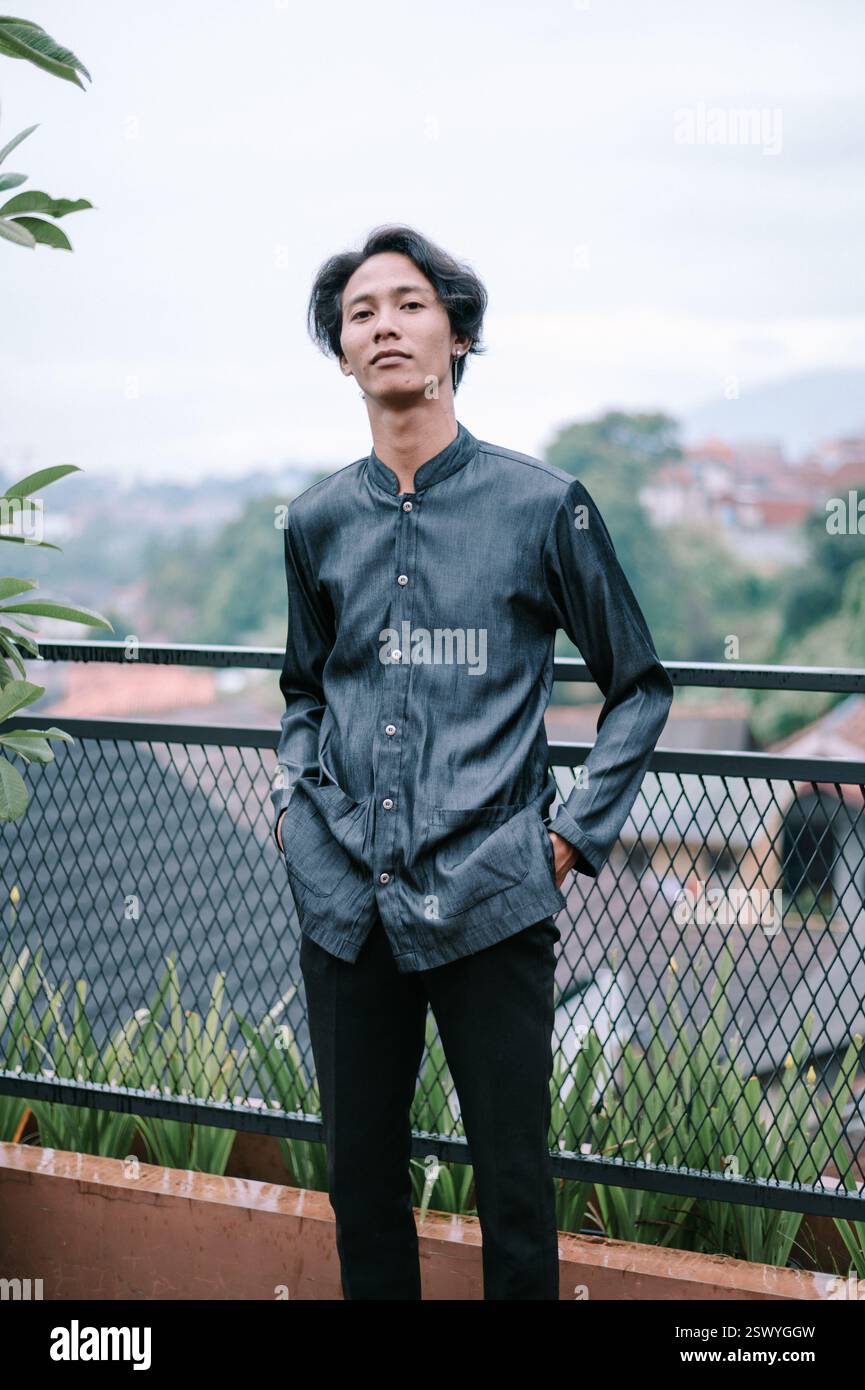 Ein junger asiatischer Mann in lässigem Hemd und schwarzer Hose posiert draußen mit den Händen in den Taschen. Der Hintergrund zeigt eine Stadt am Hang cr Stockfoto