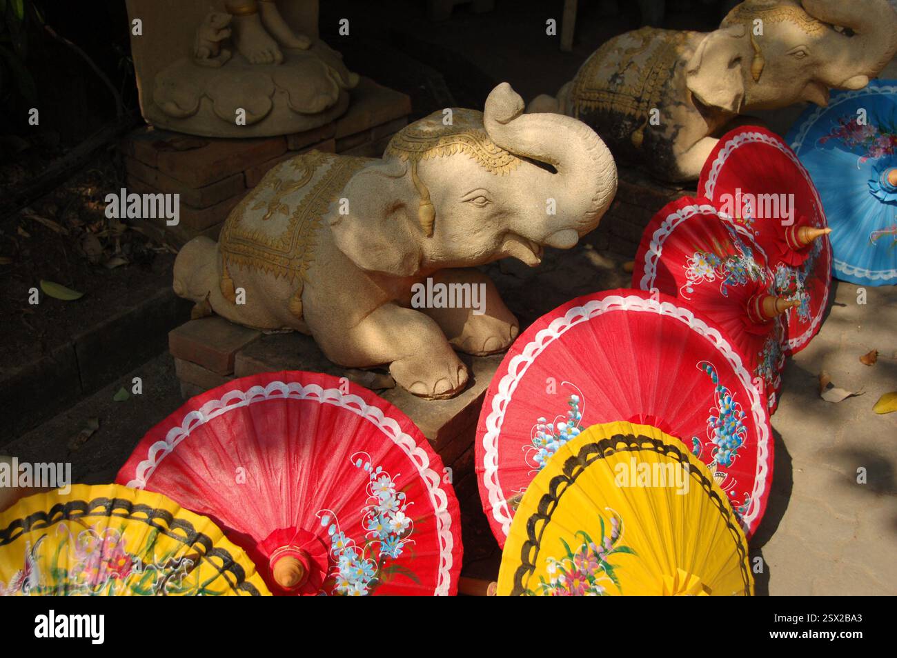 Handbemalte thailändische Papierschirme mit lebhaften Blumenmustern ruhen auf dem Boden, mit einer kleinen, hellen Steinelefanten-Statue im Hintergrund Stockfoto