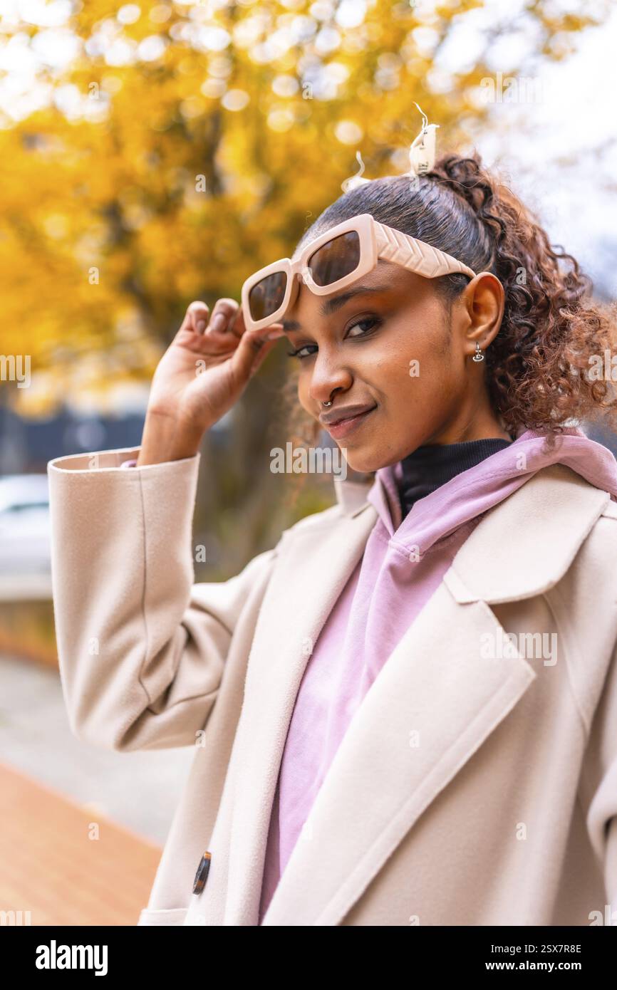 Coole afroamerikanerin, die im Herbst eine Sonnenbrille hält, um die Kamera im Mantel in der Stadt anzuschauen Stockfoto Coole afroamerikanerin, die im Herbst eine Sonnenbrille hält, um die Kamera im Mantel in der Stadt anzuschauen Stockfoto